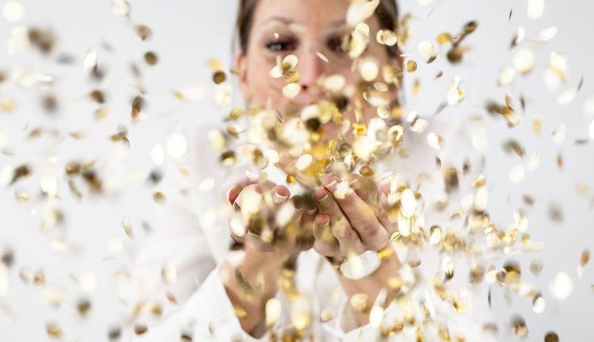 A woman' tosses gold confetti held in her cupped hands into the air, obscuring her face.